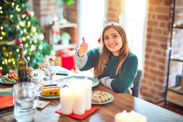Fiatal, gyönyörű nő ül és eszik a karácsonyfa körül otthon, boldog hüvelykujjával felemeli a kezét. Jóváhagyása kifejezés a kamerába mutató siker. - Fotó, kép
