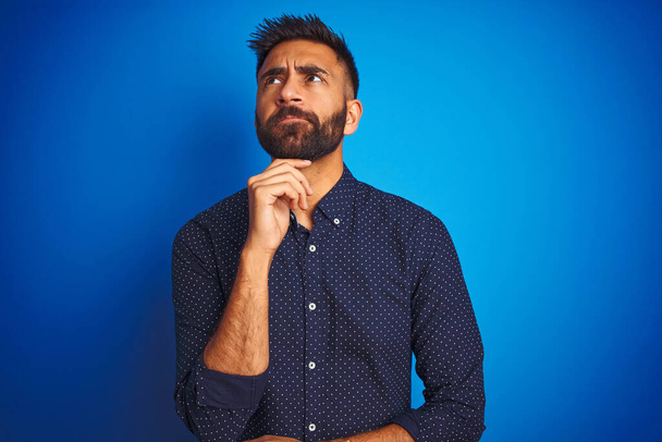 Young indian elegant man wearing shirt standing over isolated blue background with hand on chin thinking about question, pensive expression. Smiling with thoughtful face. Doubt concept. - Photo, Image
