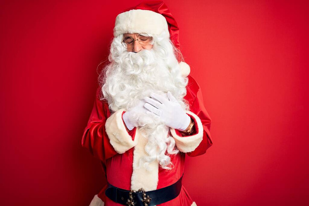 Orta yaşlı yakışıklı bir adam, Noel Baba kostümü giymiş, izole edilmiş kırmızı arka plan üzerinde duruyor, elleri göğsünde, gözleri kapalı ve yüzünde minnet dolu bir jest ile gülümsüyor. Sağlık kavramı. - Fotoğraf, Görsel