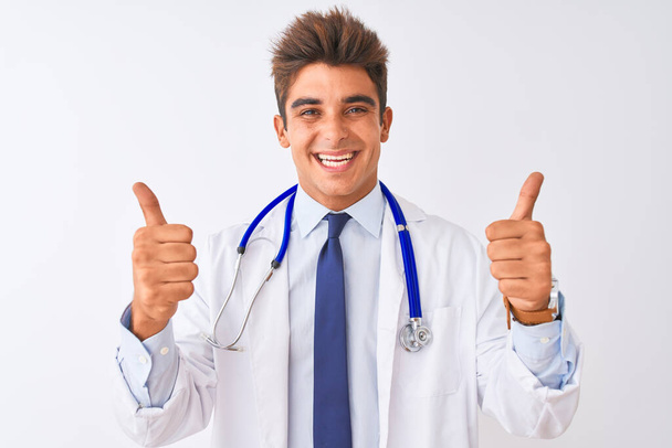 Mladý pohledný lékař se stetoskopem přes osamělé bílé pozadí znamení úspěchu dělá pozitivní gesto rukou, palce nahoru s úsměvem a šťastný. Veselý výraz a gesto vítěze. - Fotografie, Obrázek