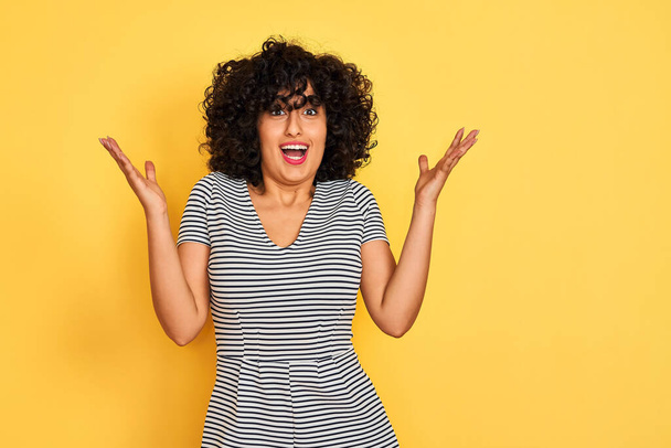 Young arab woman with curly hair wearing striped dress over isolated yellow background celebrating crazy and amazed for success with arms raised and open eyes screaming excited. Winner concept - 写真・画像