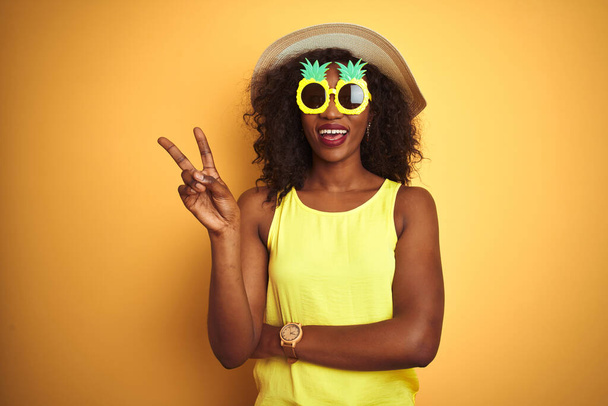 African american woman wearing funny pineapple sunglasses over isolated yellow background smiling with happy face winking at the camera doing victory sign. Number two. - 写真・画像