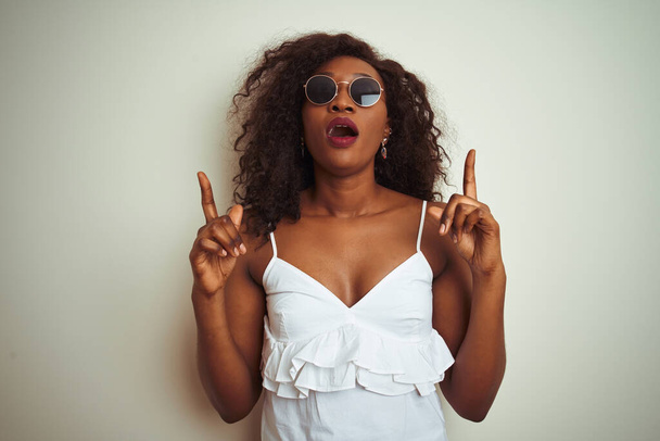 Молода афроамериканська жінка, одягнена в футболку і сонячні окуляри над ізольованим білим фоном, здивований і здивований дивлячись вгору і вказуючи пальцями і піднятими руками. - Фото, зображення