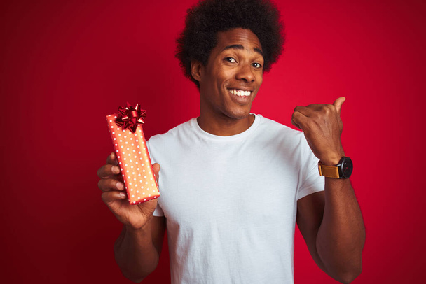 Νεαρός Αφροαμερικάνος που κρατά δώρο Αγίου Βαλεντίνου στέκεται πάνω από το απομονωμένο κόκκινο φόντο δείχνοντας και δείχνοντας με τον αντίχειρα μέχρι το πλάι με χαρούμενο πρόσωπο χαμογελώντας - Φωτογραφία, εικόνα