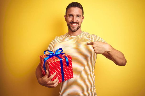 junger Mann hält ein Geburtstagsgeschenk über gelbem Hintergrund mit überraschendem Gesicht und zeigt mit dem Finger auf sich selbst - Foto, Bild
