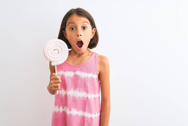 mooi kind meisje eten zoete lolly staande over geïsoleerde witte achtergrond bang in shock met een verrassing gezicht, bang en opgewonden met angst uitdrukking - Foto, afbeelding