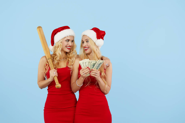 Bad Santa. Young women tweens in Santa hats are holding a bat and banknotes of money on a blue isolated background. Girls hooligans prepare for New Year's sales, robbed a bank - Foto, immagini