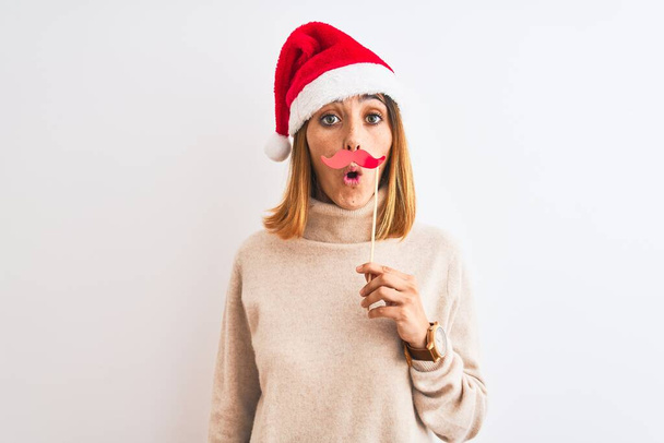 schöne rothaarige Frau trägt Weihnachtsmütze mit vorgetäuschtem Schnurrbart über isoliertem Hintergrund erschrocken mit einem Überraschungsgesicht, ängstlich und aufgeregt mit Angstausdruck - Foto, Bild