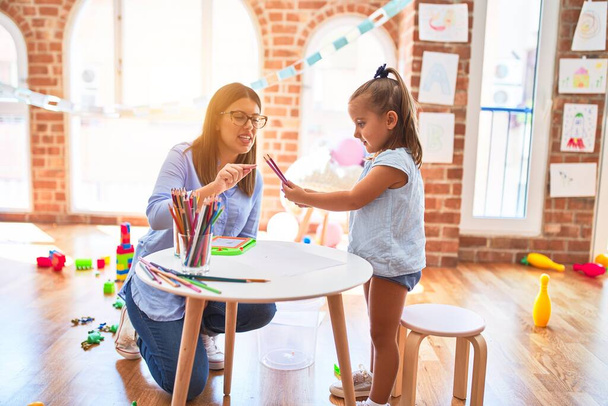 Caucasian girl kid playing and learning at playschool with female teacher. Mother and daughter at playroom around toys drawing with pencil colors - 写真・画像