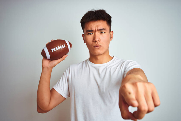 Giovane sportivo cinese asiatico che tiene la palla da rugby in piedi sopra isolato sfondo bianco che punta con il dito alla fotocamera e a voi, segno della mano, gesto positivo e fiducioso dal davanti
 - Foto, immagini