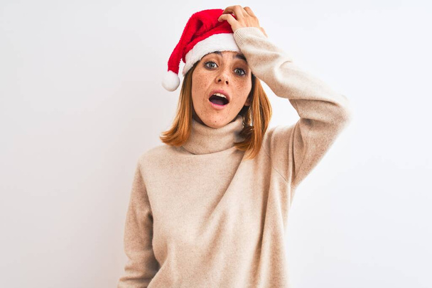 Beautiful redhead woman wearing christmas hat over isolated background surprised with hand on head for mistake, remember error. Forgot, bad memory concept. - 写真・画像