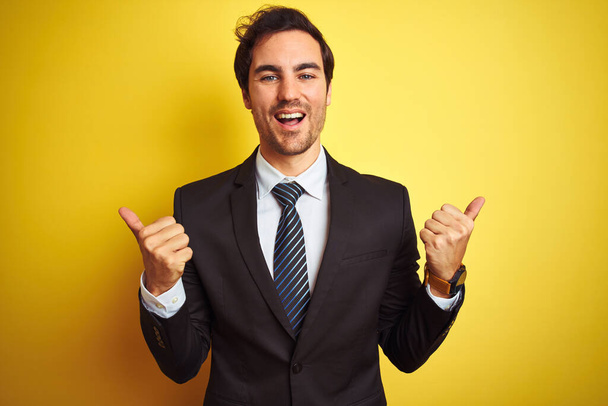 Young handsome businessman wearing suit and tie standing over isolated yellow background success sign doing positive gesture with hand, thumbs up smiling and happy. Cheerful expression and winner gesture. - Foto, immagini