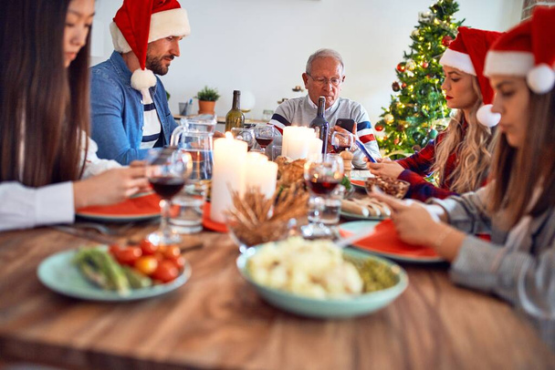 Piękna rodzina w kapeluszu Świętego Mikołaja, uśmiechnięta szczęśliwa i pewna siebie. Jedzenie pieczonego indyka przy użyciu smartfona świętującego Boże Narodzenie w domu - Zdjęcie, obraz
