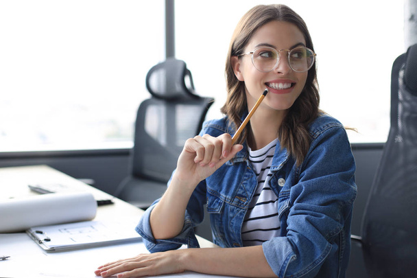 Gyönyörű fiatal nő néz kamera és kezében ceruza, miközben ül a modern irodában. - Fotó, kép