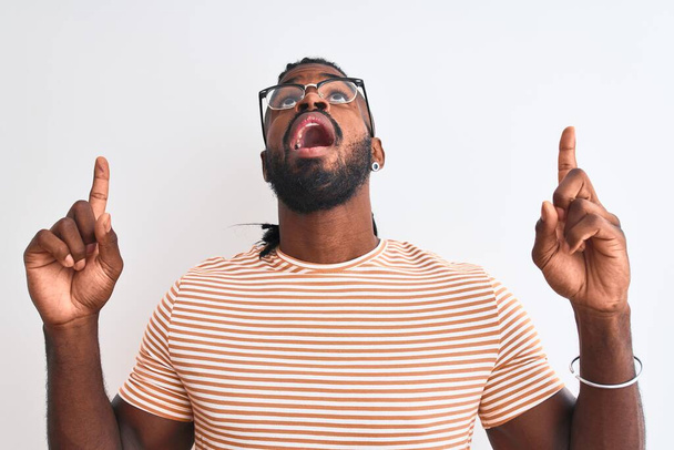African American Man ubrany w paski t-shirt i okulary na białym tle zdumiony i zaskoczony patrząc i wskazując palcami i podniesionymi ramionami. - Zdjęcie, obraz