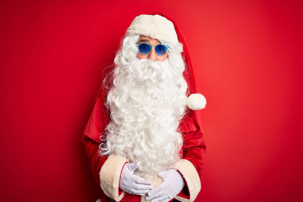 Noel Baba kostümü ve güneş gözlüğü takan orta yaşlı bir adam kırmızı arka planda korkmuş ve şaşırmış bir ifadeyle, korkuyla ve heyecanlı bir yüzle. - Fotoğraf, Görsel