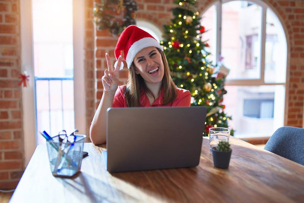 クリスマスにサンタの帽子をかぶってノートパソコンで作業しているテーブルに座っている美しい女性は、カメラが勝利サインをしているのを喜んで顔をウィンクして笑っています。第二番. - 写真・画像