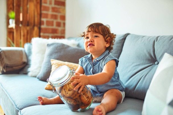 Beautiful toddler child girl holding jar of cookies sitting on the sofa - Zdjęcie, obraz