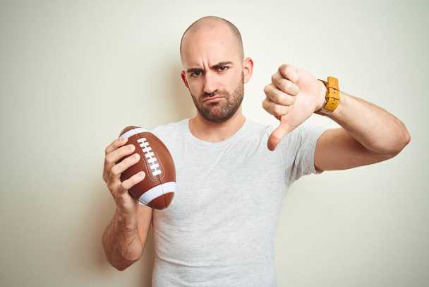 Jeune homme tenant ballon de football américain rugby sur fond isolé avec le visage en colère, signe négatif montrant aversion avec les pouces vers le bas, concept de rejet
 - Photo, image