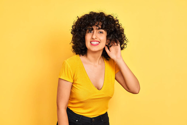 Nuori arabinainen, jolla on kiharat hiukset ja t-paita, seisoo eristetyn keltaisen taustan päällä hymyillen käsi korvan päällä kuunnellen huhuja tai juoruja. Kuurous-käsite
. - Valokuva, kuva