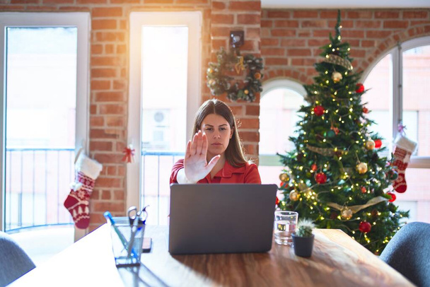 Beautiful woman sitting at the table working with laptop at home around christmas tree doing stop sing with palm of the hand. Warning expression with negative and serious gesture on the face. - 写真・画像