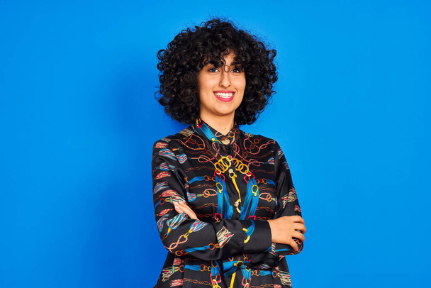 Mujer árabe joven con el pelo rizado con camisa de colores sobre fondo azul aislado cara feliz sonriendo con los brazos cruzados mirando a la cámara. Persona positiva
. - Foto, imagen