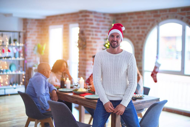 Young handsome man smiling happy and confident. Standing wearing santa hat celebrating Christmas with family at home - Photo, Image