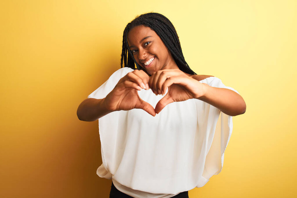 白いTシャツを着た若いアフリカ系アメリカ人女性は、ハートのシンボルと手で形を示す愛に笑みを浮かべて孤立した黄色の背景の上に立っています。ロマン主義. - 写真・画像