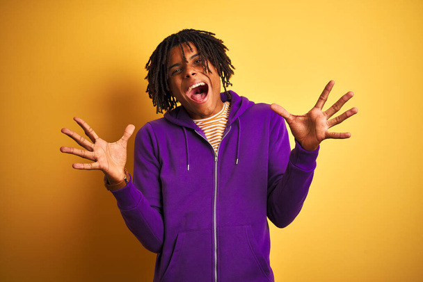 Hombre afroamericano con rastas vistiendo sudadera púrpura sobre fondo púrpura aislado celebrando loco y sorprendido por el éxito con los brazos levantados y los ojos abiertos gritando emocionados. Concepto ganador
 - Foto, imagen