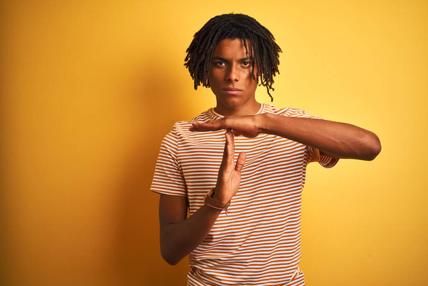 Afro człowiek z dredami w paski t-shirt stoi na odizolowanym żółtym tle Wykonywanie czasu gest z rąk, sfrustrowany i poważny twarz - Zdjęcie, obraz