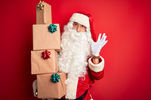 Senior άνθρωπος φορώντας κοστούμι Άγιος Βασίλης κρατώντας πύργο των δώρων πάνω από απομονωμένο κόκκινο φόντο πολύ χαρούμενος και ενθουσιασμένος, νικητής έκφραση γιορτάζει τη νίκη ουρλιάζοντας με μεγάλο χαμόγελο και σήκωσε τα χέρια - Φωτογραφία, εικόνα