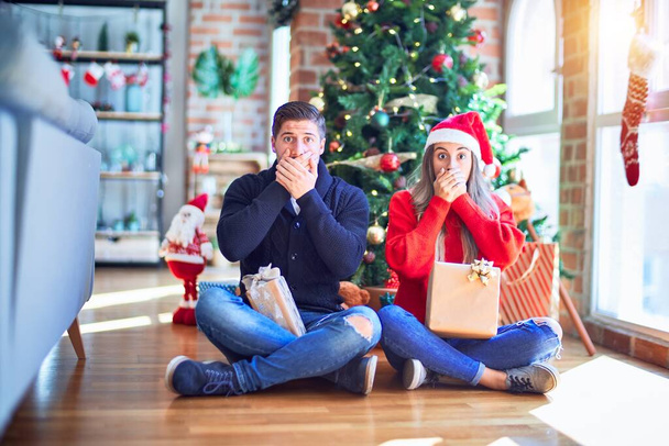 Young couple wearing santa claus hat sitting on the floor around christmas tree at home shocked covering mouth with hands for mistake. Secret concept. - Photo, image