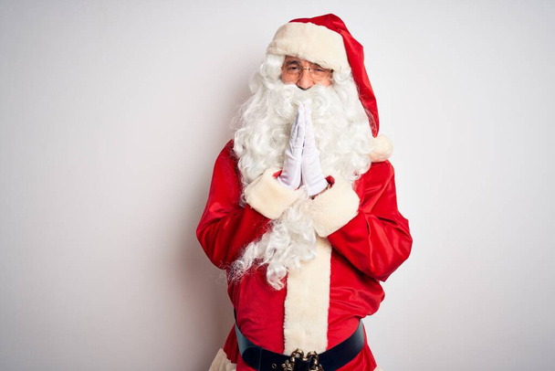 gutaussehender Mann mittleren Alters im Weihnachtsmannkostüm, der vor isoliertem weißen Hintergrund steht und mit den Händen zusammen betet und um Vergebung bittet, lächelt selbstbewusst. - Foto, Bild