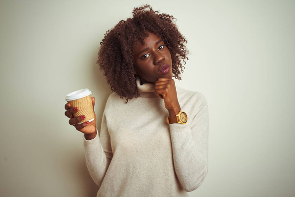 Mujer afro africana joven sosteniendo taza de café de pie sobre fondo blanco aislado cara seria pensando en la pregunta, idea muy confusa
 - Foto, imagen
