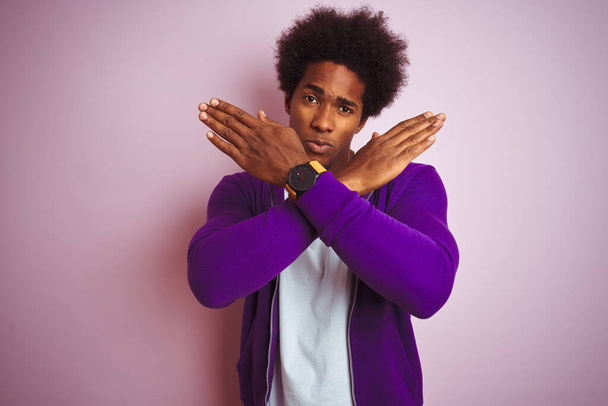 若いアフリカ系アメリカ人男性が紫のトレーナーを着てピンクの背景の上に立って否定的な記号を行う腕を横断拒否式,怒っている顔 - 写真・画像