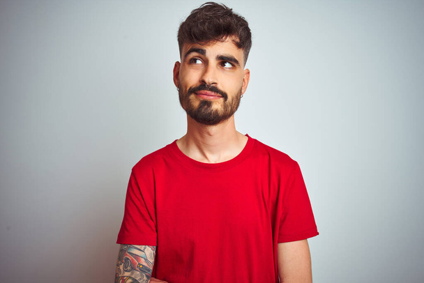 Young man with tattoo wearing red t-shirt standing over isolated white background smiling looking to the side and staring away thinking. - Foto, imagen