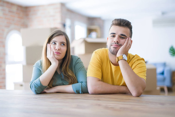 Young couple sitting on the table movinto to new home with carboard boxes behind them thinking looking tired and bored with depression problems with crossed arms. - Foto, Imagen