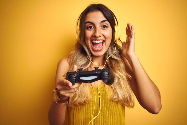 Young beautiful woman playing video game using gamepad over yellow isolated background very happy and excited, winner expression celebrating victory screaming with big smile and raised hands - Photo, image