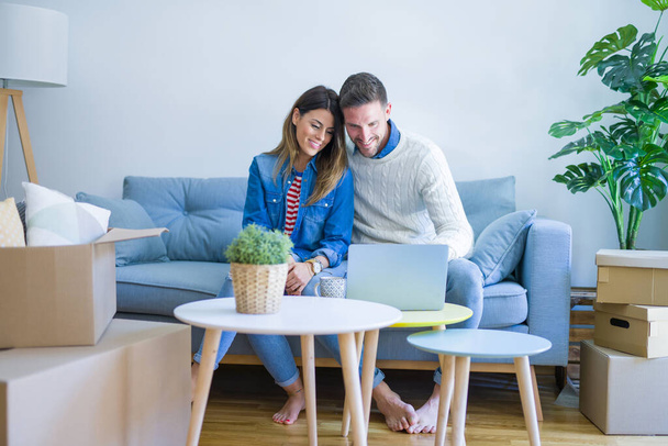 Young beautiful couple sitting on the sofa drinking cup of coffee using laptop at new home around cardboard boxes - Photo, image
