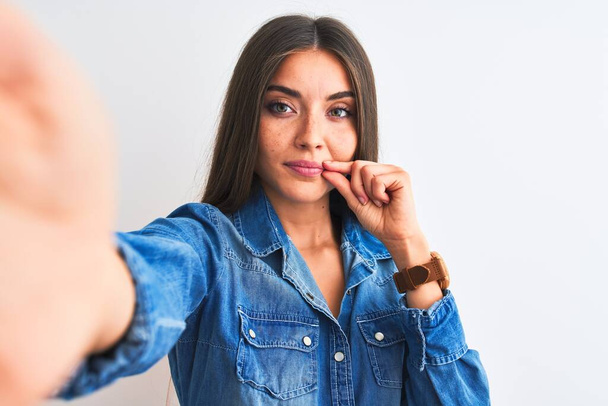 Krásná žena v džínové košili, aby selfie kamerou přes izolované bílé pozadí ústa a rty zavřené jako zip s prsty. Tajné a tiché, tabu mluvení - Fotografie, Obrázek