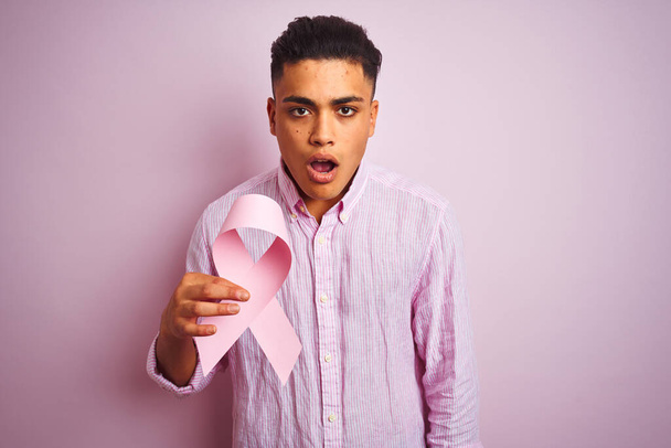 Joven brasileño sosteniendo cinta de cáncer parado sobre fondo rosa aislado asustado en shock con una cara sorpresa, asustado y excitado con expresión de miedo
 - Foto, Imagen