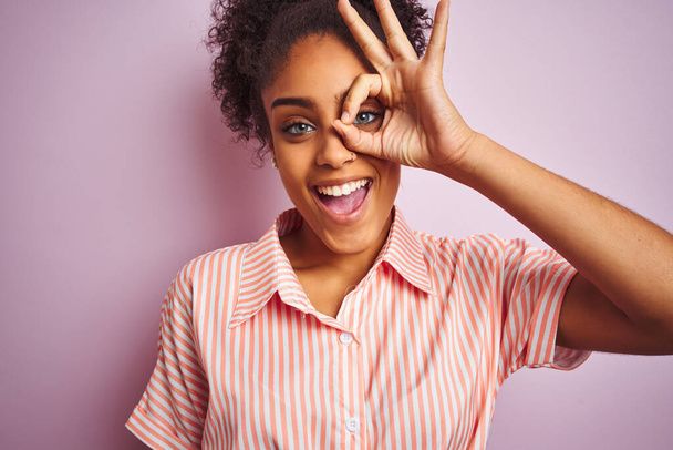 Femme afro-américaine portant une chemise rayée décontractée debout sur un fond rose isolé avec un visage heureux souriant faisant signe ok avec la main sur les yeux regardant à travers les doigts
 - Photo, image