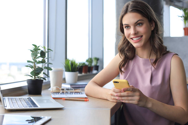 Beautiful business woman is using her smartphone with smile while sitting at her working place - Foto, Bild