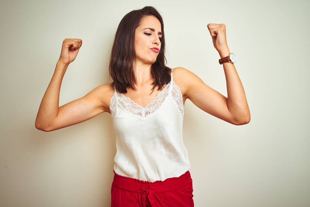 Young beautiful woman wearing t-shirt standing over white isolated background showing arms muscles smiling proud. Fitness concept. - Zdjęcie, obraz