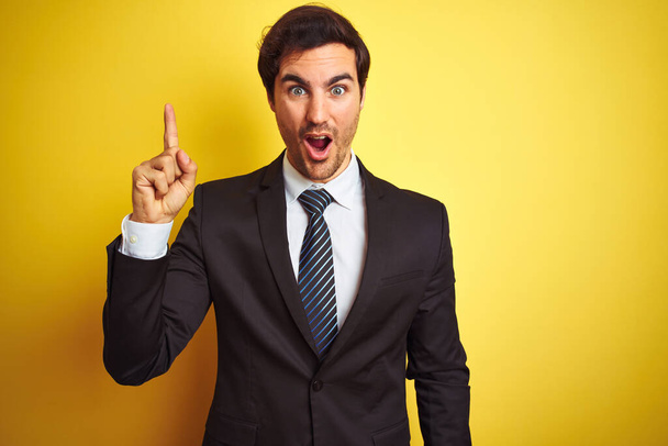 Young handsome businessman wearing suit and tie standing over isolated yellow background pointing finger up with successful idea. Exited and happy. Number one. - Fotoğraf, Görsel