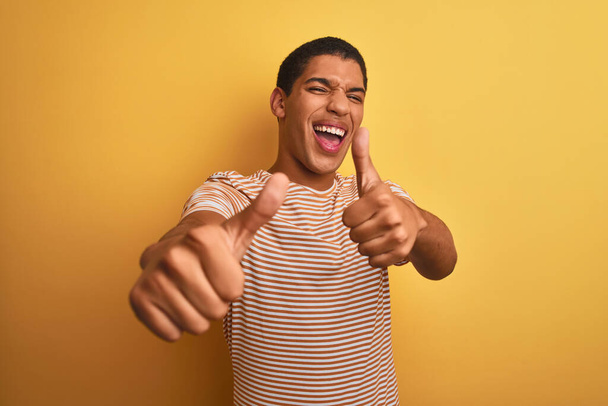 Jonge knappe arabische man met gestreept t-shirt over geïsoleerde gele achtergrond die positief gebaar met de hand, duimen omhoog lachend en blij voor succes goedkeurt. Winnaar gebaar. - Foto, afbeelding