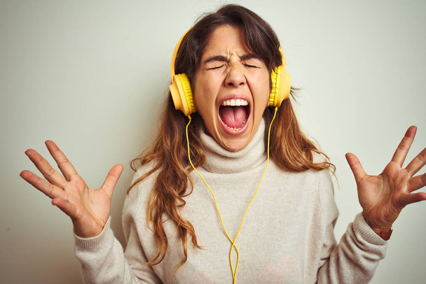 Young beautiful woman listening to music using headphones over white isolated background celebrating mad and crazy for success with arms raised and closed eyes screaming excited. Winner concept - Foto, Imagen