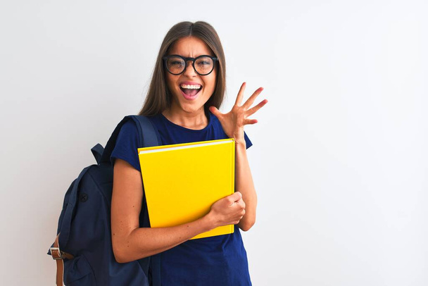 Young student woman wearing backpack glasses holding book over isolated white background very happy and excited, winner expression celebrating victory screaming with big smile and raised hands - Photo, Image