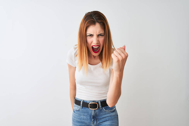 Beautiful redhead woman wearing casual t-shirt standing over isolated white background angry and mad raising fist frustrated and furious while shouting with anger. Rage and aggressive concept. - Photo, Image