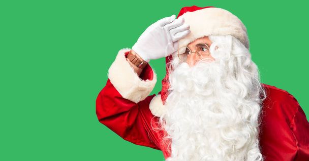 Middle age handsome man wearing Santa Claus costume and beard standing very happy and smiling looking far away with hand over head. Searching concept. - Photo, Image